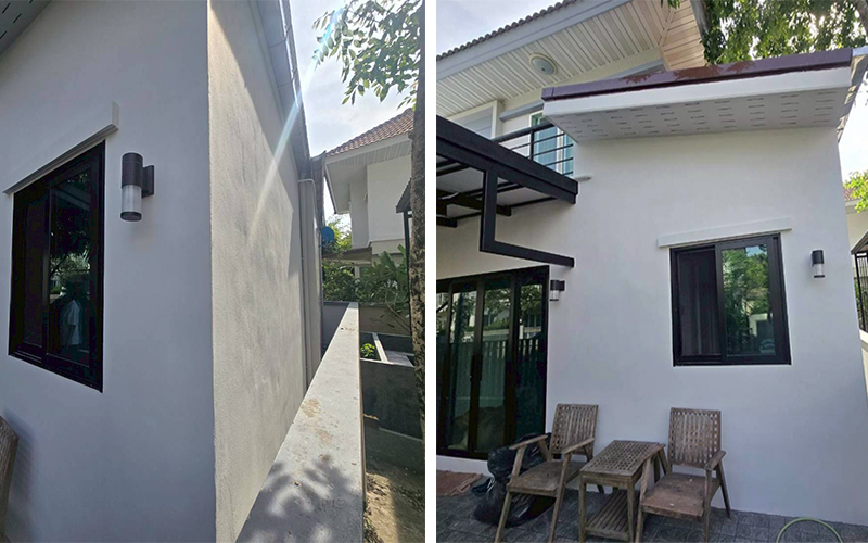 ผนังฉาบปูนเรียบสีขาว ติดตั้งหน้าต่างอลูมิเนียมกรอบดำ พร้อมโคมไฟผนัง และระเบียงชั้น 2